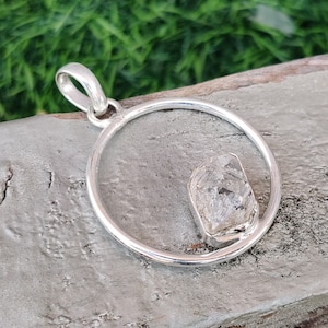 May include: A silver pendant with a clear, faceted crystal set in a circle. The pendant is on a wooden surface.