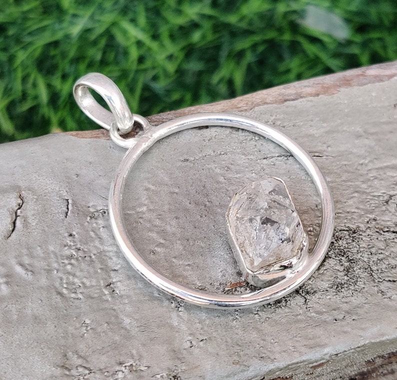 May include: A silver pendant with a clear, faceted crystal set in a circle. The pendant is on a wooden surface.