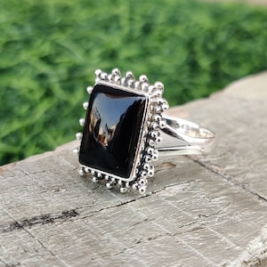 Könnte beinhalten: Ein silberner Ring mit einem schwarzen quadratischen Edelstein, der in einem dekorativen Rahmen gefasst ist. Der Ring liegt auf einer Holzoberfläche.