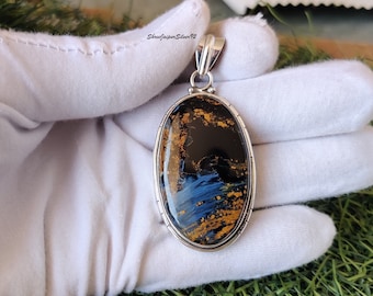 Puede incluir: Un colgante de plata con una piedra grande ovalada de ojo de tigre azul y dorado. El colgante tiene una anilla de plata y está engastado en un engaste de plata.