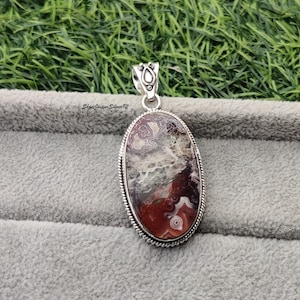Puede incluir: Un colgante de plata con una piedra de ágata de forma ovalada. La piedra tiene un patrón marrón, blanco y gris. El colgante está sobre un fondo gris.