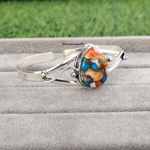 May include: A silver bangle bracelet with a teardrop-shaped turquoise stone set in the center. The stone is a mix of blue, green, orange, and white.