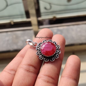 May include: A silver pendant with a red ruby gemstone set in a floral design. The pendant is held in a hand.