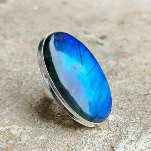 Könnte beinhalten: Ein silberner Ring mit einem großen ovalen blauen Labradorit-Edelstein. Der Stein hat einen irisierenden Glanz und zeigt eine Reihe von blauen und grünen Farbtönen.
