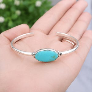 May include: A silver cuff bracelet featuring a central oval-shaped turquoise stone. The polished silver band has an open design, and the turquoise stone displays a bright blue colour with subtle natural brown matrix inclusions in the centre.