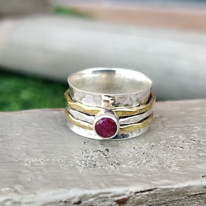 May include: A silver and gold-tone spinner ring with a hammered texture. The ring features a central, round, deep red gemstone. The design includes multiple bands, with a rotating outer band for a fidget-friendly feature.