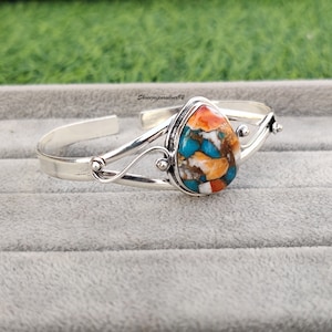 May include: A silver bangle bracelet with a teardrop-shaped turquoise stone set in the center. The stone is a mix of blue, green, orange, and white.
