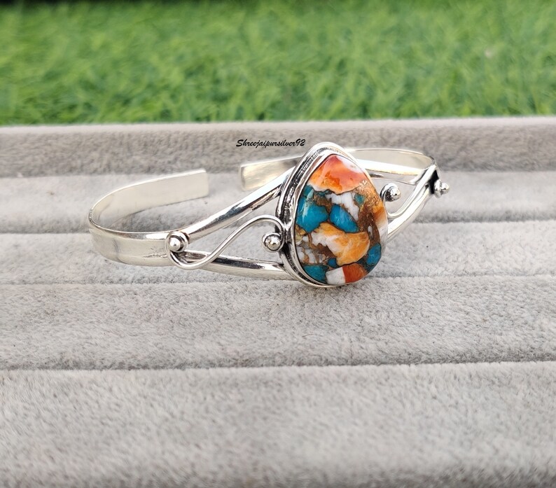 May include: A silver bangle bracelet with a teardrop-shaped turquoise stone set in the center. The stone is a mix of blue, green, orange, and white.