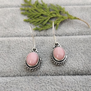 Può includere: Un paio di orecchini pendenti in argento con pietre preziose ovali rosa chiaro. Le pietre sono incastonate in un motivo a corda intrecciata. Gli orecchini sono esposti su una superficie grigia con un rametto di fogliame verde.