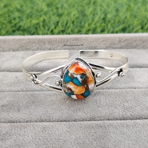 May include: A silver bangle bracelet with a teardrop-shaped turquoise, orange, and white stone set in the center.