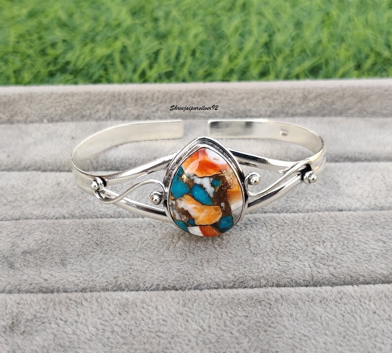 May include: A silver bangle bracelet with a teardrop-shaped turquoise, orange, and white stone set in the center.