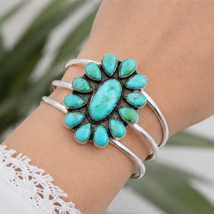 May include: A silver cuff bracelet featuring a large central oval turquoise stone surrounded by twelve smaller teardrop-shaped turquoise stones. The stones exhibit natural blue-green colour variations and dark matrix patterns set in a polished silver frame.