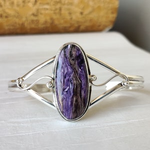 Pode incluir: Uma pulseira de prata com uma pedra preciosa ovalada de cor púrpura e cinza engastada no centro. A pulseira tem um design delicado e intrincado.