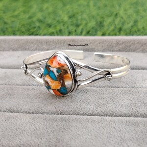 May include: A silver cuff bracelet with a teardrop-shaped turquoise stone set in the center. The stone is a mix of orange, blue, and white colors. The bracelet has a delicate, swirling design.