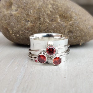 May include: A silver spinning ring with three red gemstones. The ring has a wide band and is decorated with three thin bands.