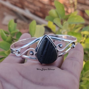以下が含まれることがあります： 黒い宝石が涙の形をしたシルバーのバングルブレスレット。ブレスレットは繊細で装飾的なデザインです。