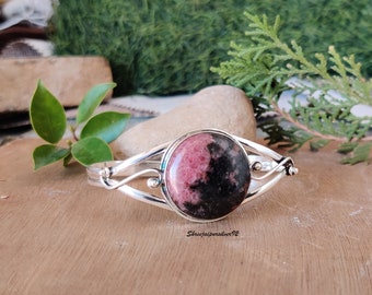 May include: A silver bangle bracelet featuring a pink and black gemstone cabochon. The bracelet has a delicate, twisted design.