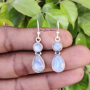 Può includere: Un paio di orecchini in argento con design a pietra di luna. Ogni orecchino presenta una piccola pietra di luna rotonda nella parte superiore e una più grande a forma di goccia nella parte inferiore. Gli orecchini sono tenuti in mano su uno sfondo di foglie verdi.