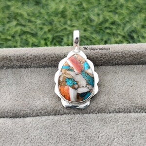 Pu&ograve; includere: Un ciondolo a forma di lacrima con una montatura in argento e un'incisione di pietra colorata. La pietra &egrave; un mix di turchese, corallo e bianco, con un motivo maculato. Il ciondolo &egrave; su uno sfondo grigio.