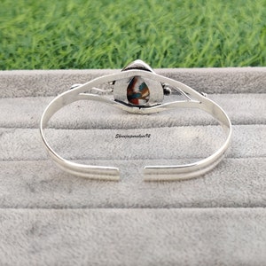 May include: Silver bangle bracelet with a colorful stone inlay. The stone is a teardrop shape and has a variety of colors, including blue, green, red, and white.
