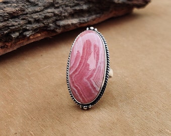 May include: A silver ring featuring a large, oval-shaped pink gemstone with white veining. The gemstone is set in a silver bezel with a twisted rope design around the edge. The ring is displayed on a wooden surface.