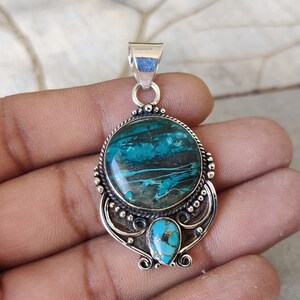 May include: A silver pendant with a large oval turquoise stone set in a silver bezel. The stone has a green and black marbled pattern. The pendant is attached to a silver bail.