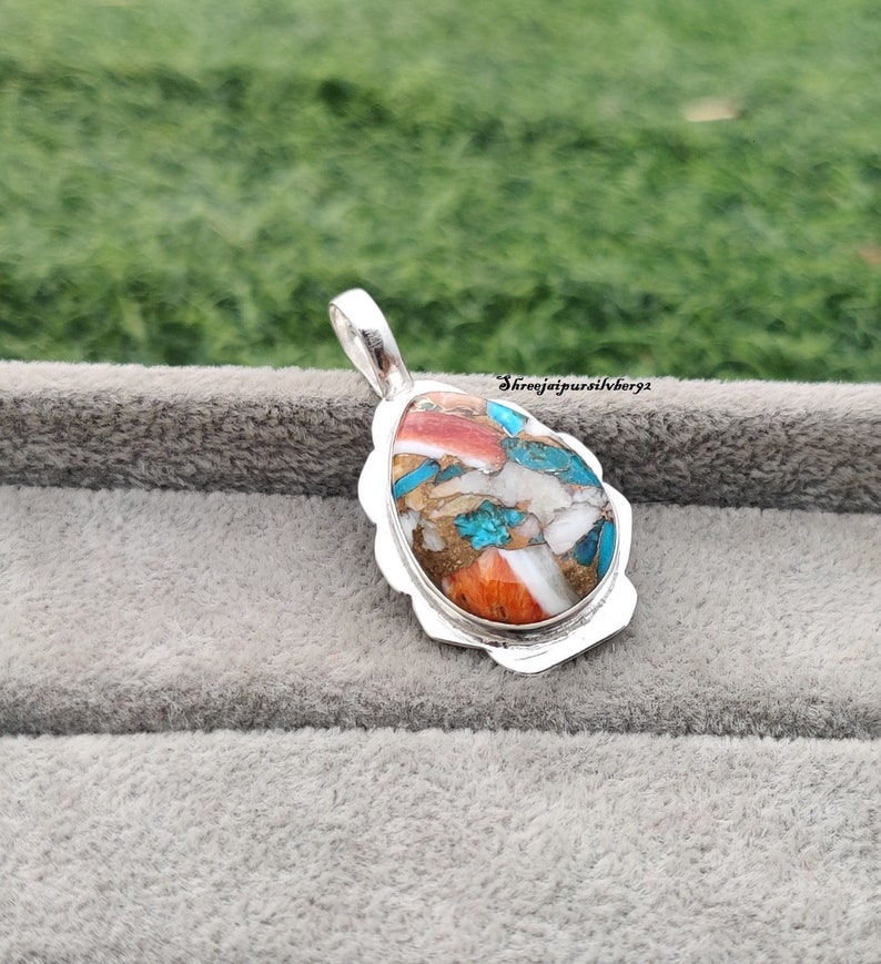 Pu&ograve; includere: Un ciondolo d'argento con un'incisione di pietra colorata. La pietra &egrave; un mix di turchese, rosso, bianco e marrone. Il ciondolo &egrave; di forma ovale e ha un'anellina d'argento.