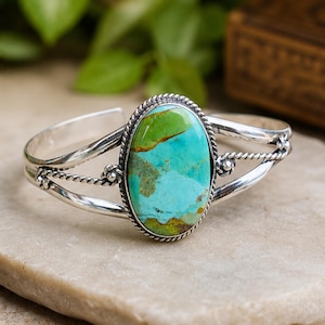 May include: A silver cuff bracelet featuring a large, oval-shaped turquoise stone with vibrant green and blue marbling. The stone is set in a twisted rope-style silver bezel, and the band consists of two polished silver wires with twisted accents.