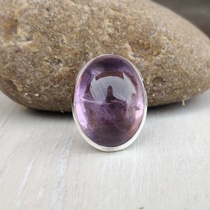 Könnte beinhalten: Ein silberner Ring mit einem großen ovalen Amethyst-Edelstein. Der Edelstein ist tiefviolett und hat einen leichten Schimmer.