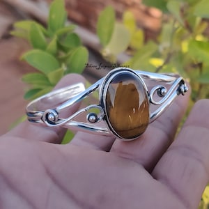 May include: Silver bangle bracelet with a large oval tiger's eye gemstone. The bracelet has a delicate, swirling design.