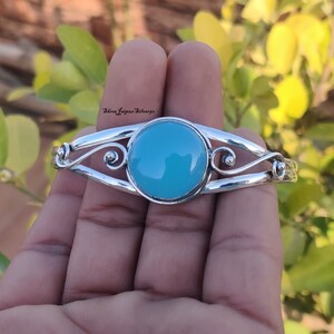 以下が含まれることがあります： ターコイズのcabochonをセットしたシルバーのバングルブレスレット。ブレスレットは、渦巻き模様と中央の石を配した繊細で装飾的なデザインです。