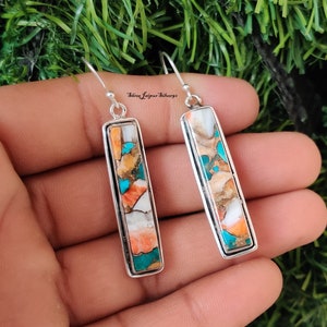 Op de afbeelding: Een paar zilveren oorhangers met rechthoekige inlegsels van turquoise, oranje en wit. De oorbellen hebben een zilveren haaksluiting.