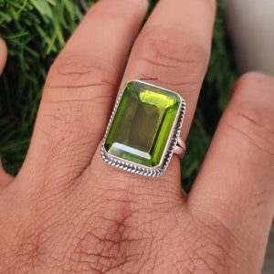 Puede incluir: Un anillo de plata con una gran piedra preciosa rectangular verde. El anillo tiene un diseño trenzado detallado alrededor de la piedra.