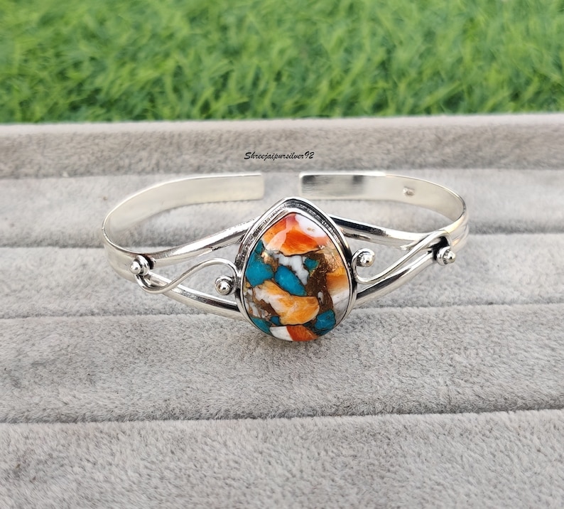 May include: A silver bangle bracelet with a teardrop-shaped turquoise, orange, and white stone set in the center.