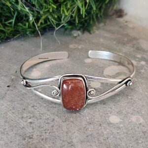 May include: Silver cuff bracelet with a rectangular, reddish-brown gemstone. The bracelet has a delicate, twisted design and is stamped "Sterling Silver".
