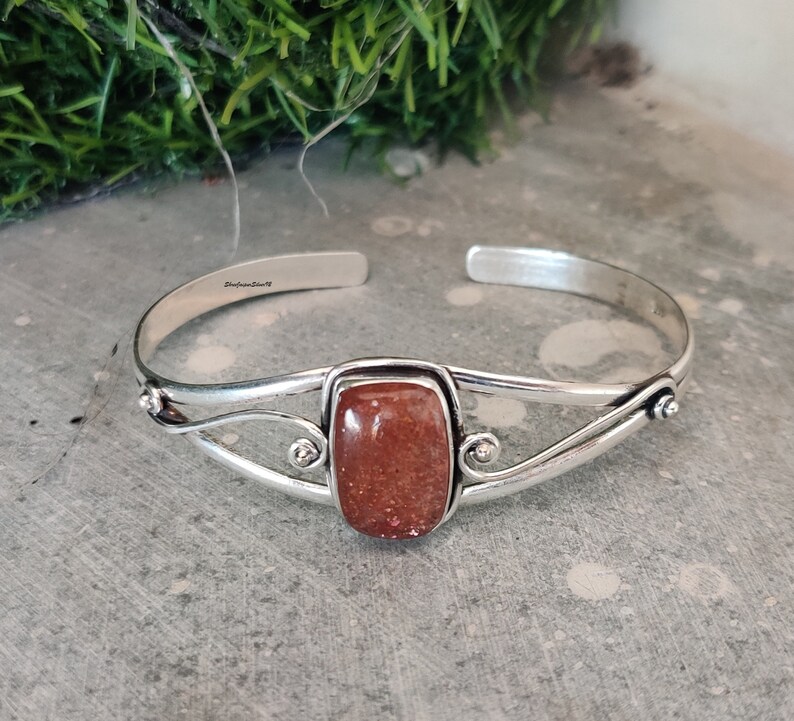 May include: Silver cuff bracelet with a rectangular, reddish-brown gemstone. The bracelet has a delicate, twisted design and is stamped "Sterling Silver".