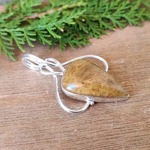 May include: A silver pendant with a teardrop-shaped clear quartz crystal with brown inclusions. The pendant is on a wooden surface.