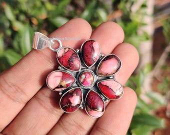 May include: A silver pendant with a flower design. The pendant features a central pink stone surrounded by eight petal-shaped stones in shades of pink and burgundy. The pendant is held in a hand against a blurred green background.