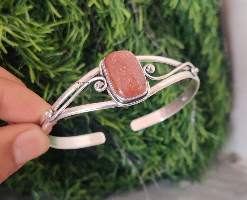 May include: A silver bangle bracelet with a rectangular, orange sunstone gemstone. The bracelet has a delicate, ornate design with swirls.