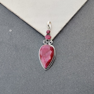 May include: A silver pendant featuring a large, teardrop-shaped ruby gemstone. The pendant is embellished with three smaller ruby gemstones.