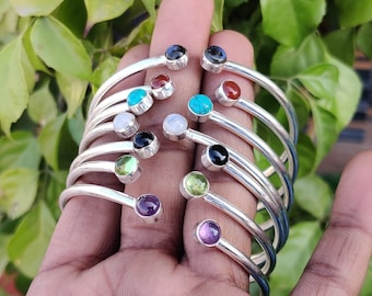 Puede incluir: Una pila de brazaletes de plata con piedras preciosas de diferentes colores. Las piedras preciosas incluyen amatista, turquesa, verde, rojo, negro y blanco.