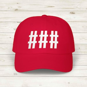 May include: A red baseball cap featuring a white hashtag symbol. The cap has a curved peak and a structured crown. The background is a light wood grain.