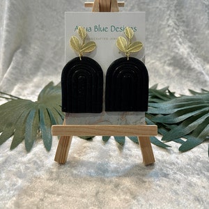 May include: Black polymer clay earrings with gold leaf accents. The earrings are shaped like arches with a gold leaf design at the top. The earrings are displayed on a wooden easel with the text "Aqua Blue Designs Handcrafted Jewelry".