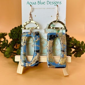 Puede incluir: Pendientes de resina dorada y azul con un arco dorado y un rectángulo dorado con un óvalo recortado. Los pendientes están hechos a mano y están etiquetados como "Aqua Blue Designs, Joyería artesanal".