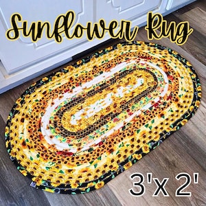 May include: An oval braided rug featuring a repeating sunflower pattern in yellow, orange, and brown tones with a black border. The rug displays the text Sunflower Rug and 3' x 2' in the image, indicating its dimensions in feet.