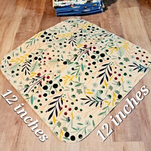 K&ouml;nnte beinhalten: Ein quadratisches Stoffk&uuml;chenhandtuch mit einer Gr&ouml;&szlig;e von 30,5 cm x 30,5 cm mit einem floralen Muster in Gr&uuml;n-, Gelb-, Rot- und Schwarzt&ouml;nen auf cremefarbenem Hintergrund.