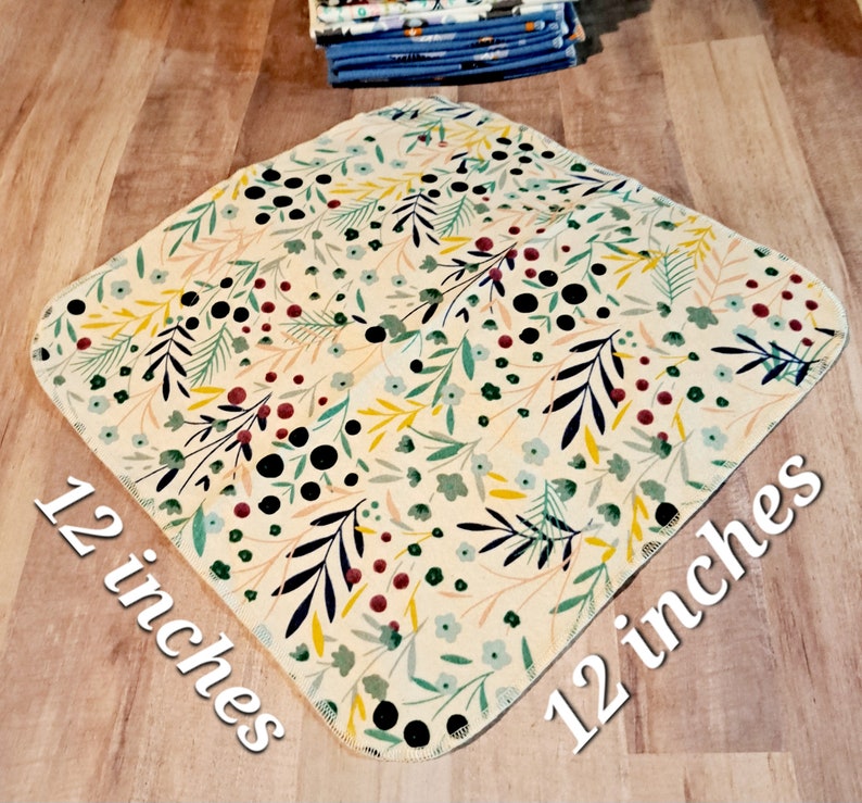 K&ouml;nnte beinhalten: Ein quadratisches Stoffk&uuml;chenhandtuch mit einer Gr&ouml;&szlig;e von 30,5 cm x 30,5 cm mit einem floralen Muster in Gr&uuml;n-, Gelb-, Rot- und Schwarzt&ouml;nen auf cremefarbenem Hintergrund.