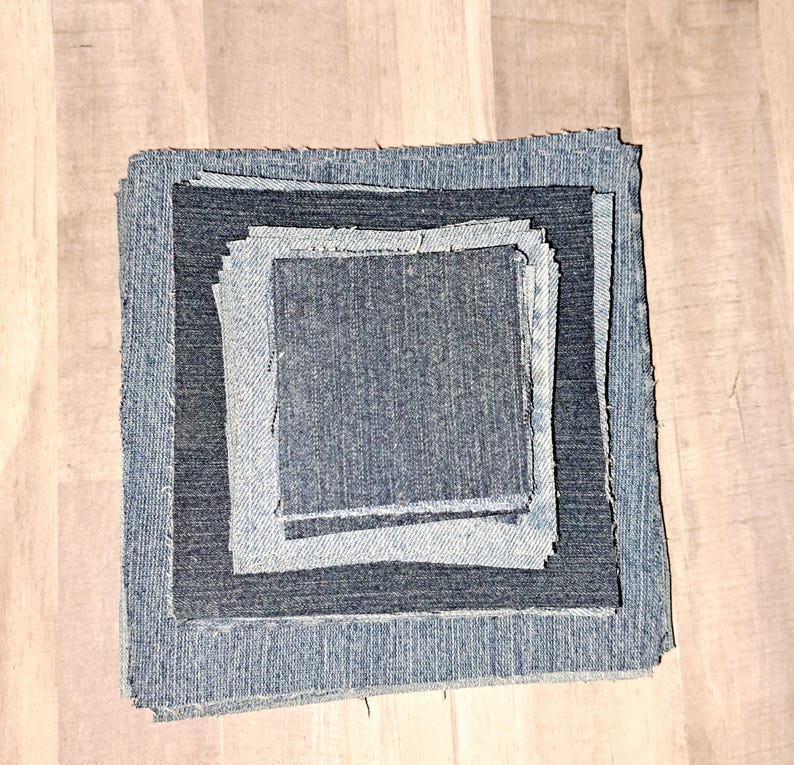 May include: Four squares of denim fabric, stacked on top of each other, with the largest square on the bottom and the smallest square on top. The fabric is a dark blue color and has a slightly textured surface.
