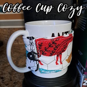 May include: A white coffee mug with a red, white, and blue fabric cup cozy. The cozy has a chicken and floral design. The text "ANTI" is visible on the mug.