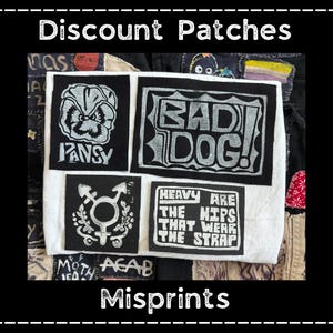 May include: A collection of black and white patches with various designs and text. The patches include a pansy flower, the words "BAD DOG!", a gender symbol, and the phrase "HEAVY ARE THE HIPS THAT WEAR THE STRAP". The text "Discount Patches" and "Misprints" are also visible.
